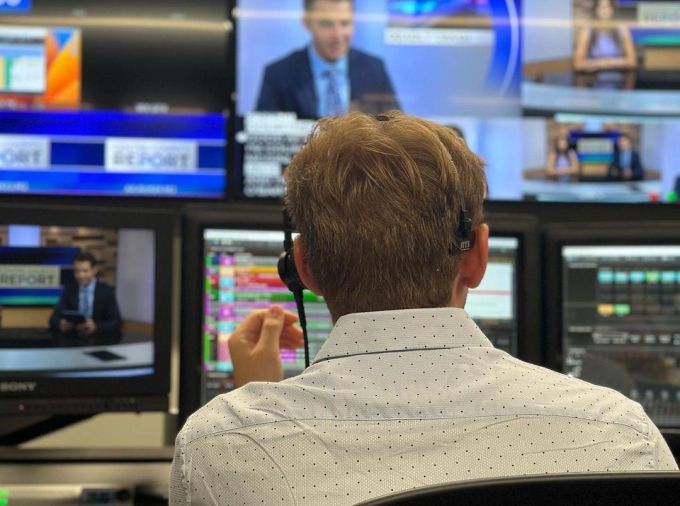 Back of a student sitting in a television studio control room producing content in the studio while watching numerous monitors