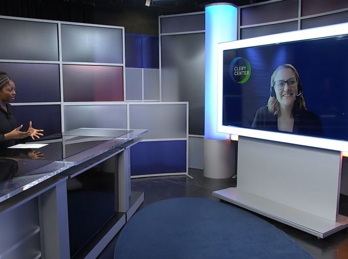A student interviewer sitting behind a news desk on set virtually interviews a woman on a large monitor