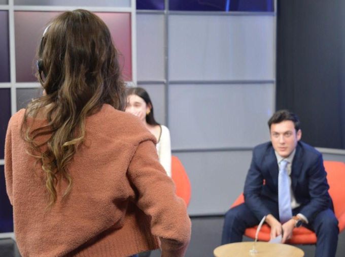 A student in a blue suit sitting on an orange chair focuses intently on instructions coming from a woman producer in the foreground in a brown sweater and jeans