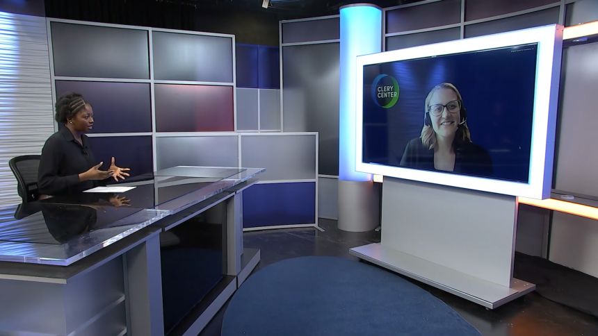 A student interviewer sitting behind a news desk on set virtually interviews a woman on a large monitor